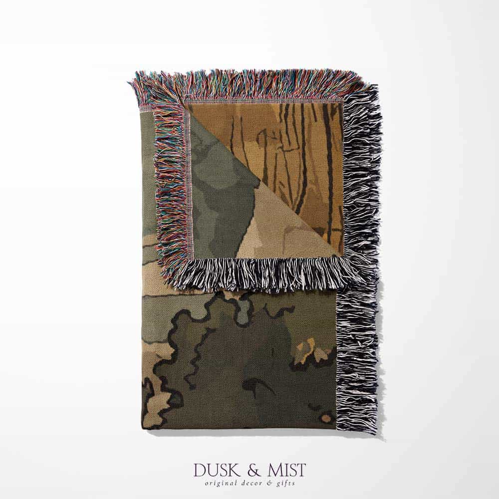 Woven cotton blanket featuring a small ghost standing in a golden meadow facing a brown and white cow, with a rustic cabin and forested hills in the background.