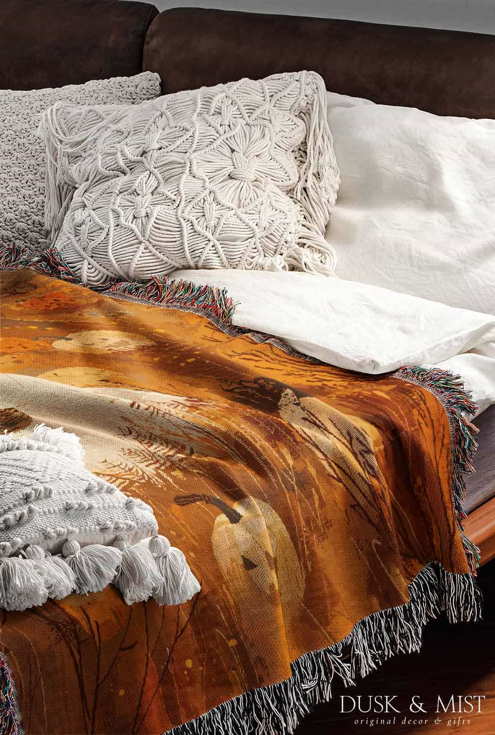 Woven cotton blanket showing a small ghost holding a basket, surrounded by pumpkins and autumn foliage in warm golden and orange tones.