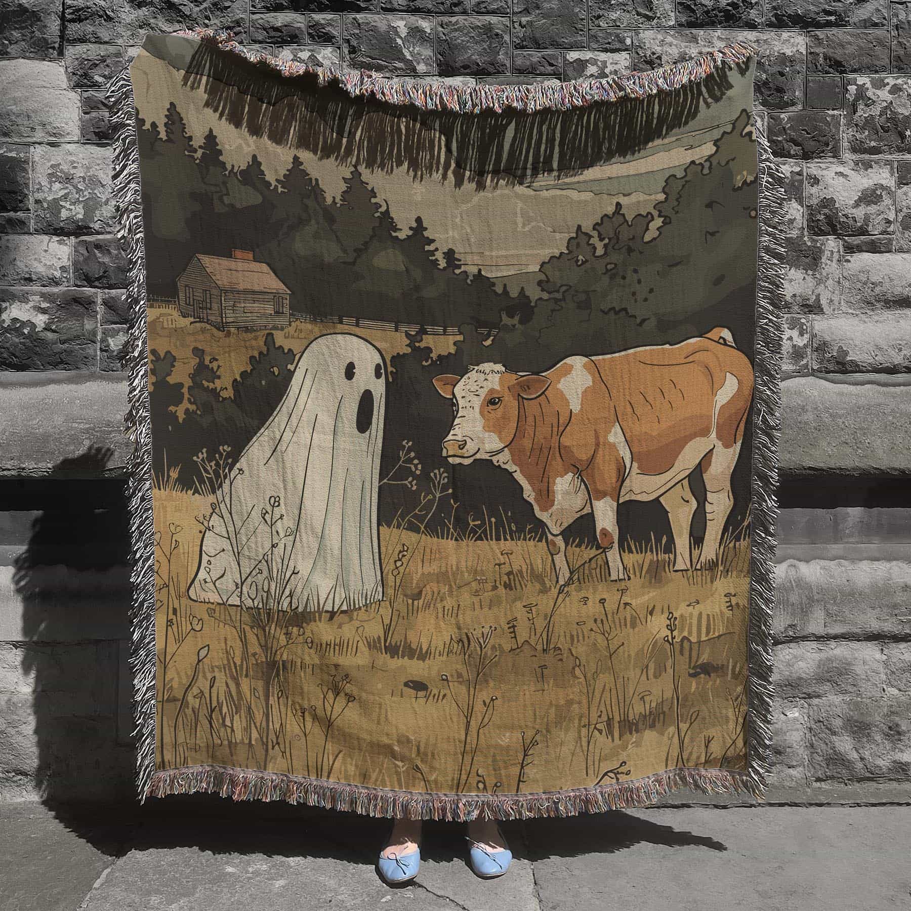 Woven cotton blanket featuring a small ghost standing in a golden meadow facing a brown and white cow, with a rustic cabin and forested hills in the background.