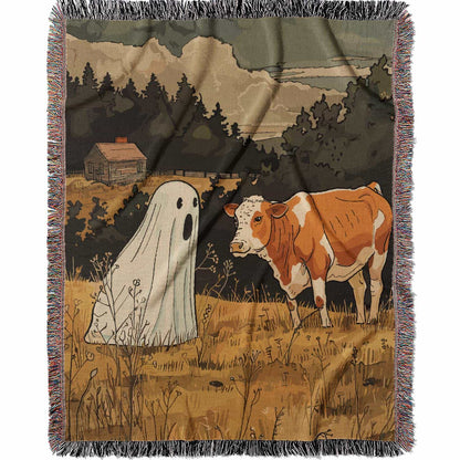 Woven cotton blanket featuring a small ghost standing in a golden meadow facing a brown and white cow, with a rustic cabin and forested hills in the background.