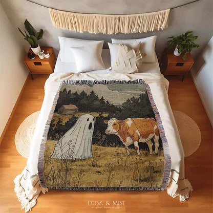 Woven cotton blanket featuring a small ghost standing in a golden meadow facing a brown and white cow, with a rustic cabin and forested hills in the background.