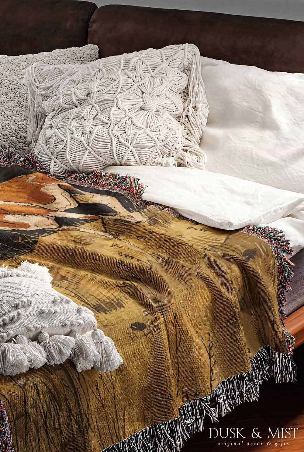 Woven cotton blanket featuring a small ghost standing in a golden meadow facing a brown and white cow, with a rustic cabin and forested hills in the background.