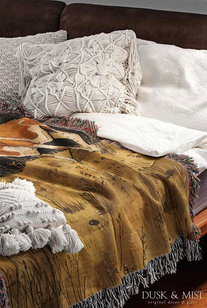 Woven cotton blanket featuring a small ghost standing in a golden meadow facing a brown and white cow, with a rustic cabin and forested hills in the background.