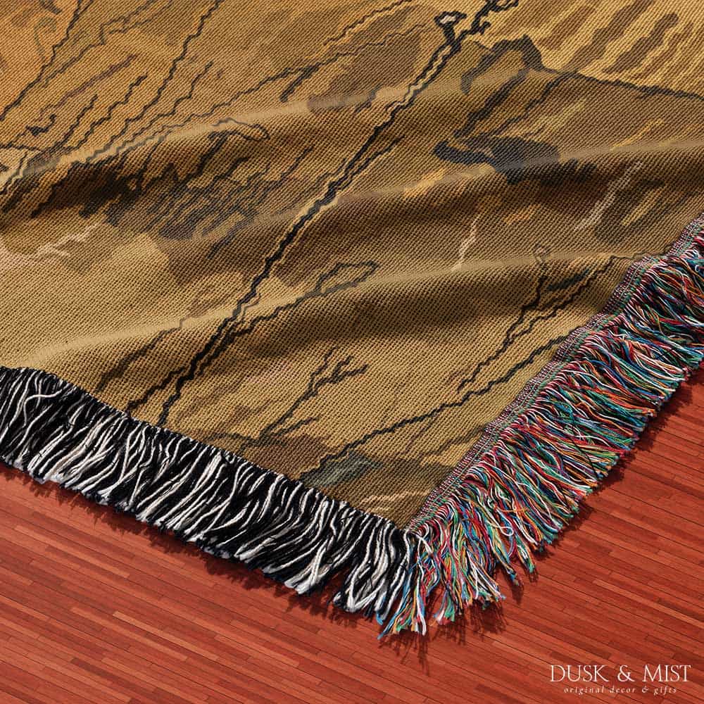 Woven cotton blanket featuring a small ghost standing in a golden meadow facing a brown and white cow, with a rustic cabin and forested hills in the background.