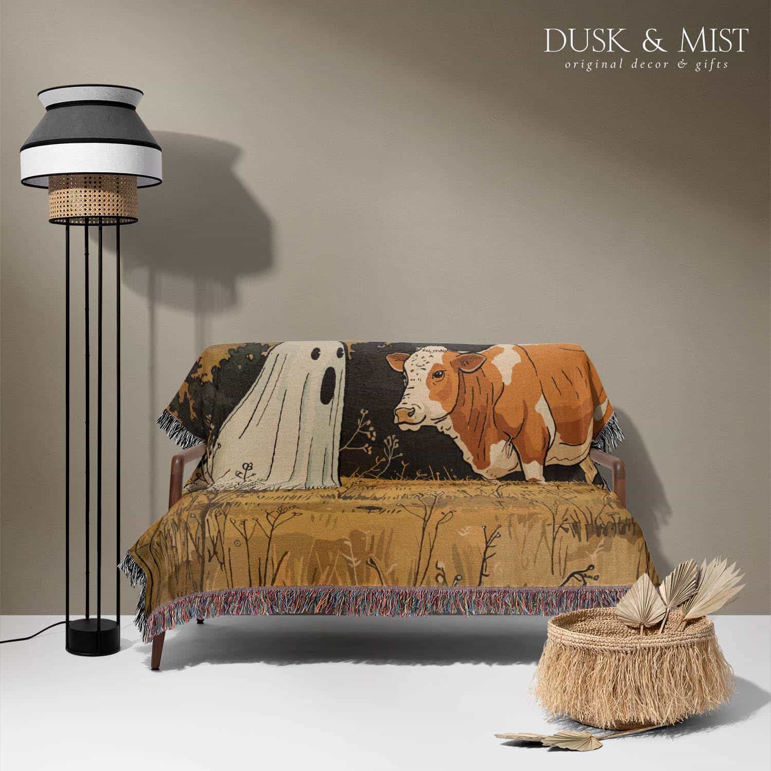 Woven cotton blanket featuring a small ghost standing in a golden meadow facing a brown and white cow, with a rustic cabin and forested hills in the background.