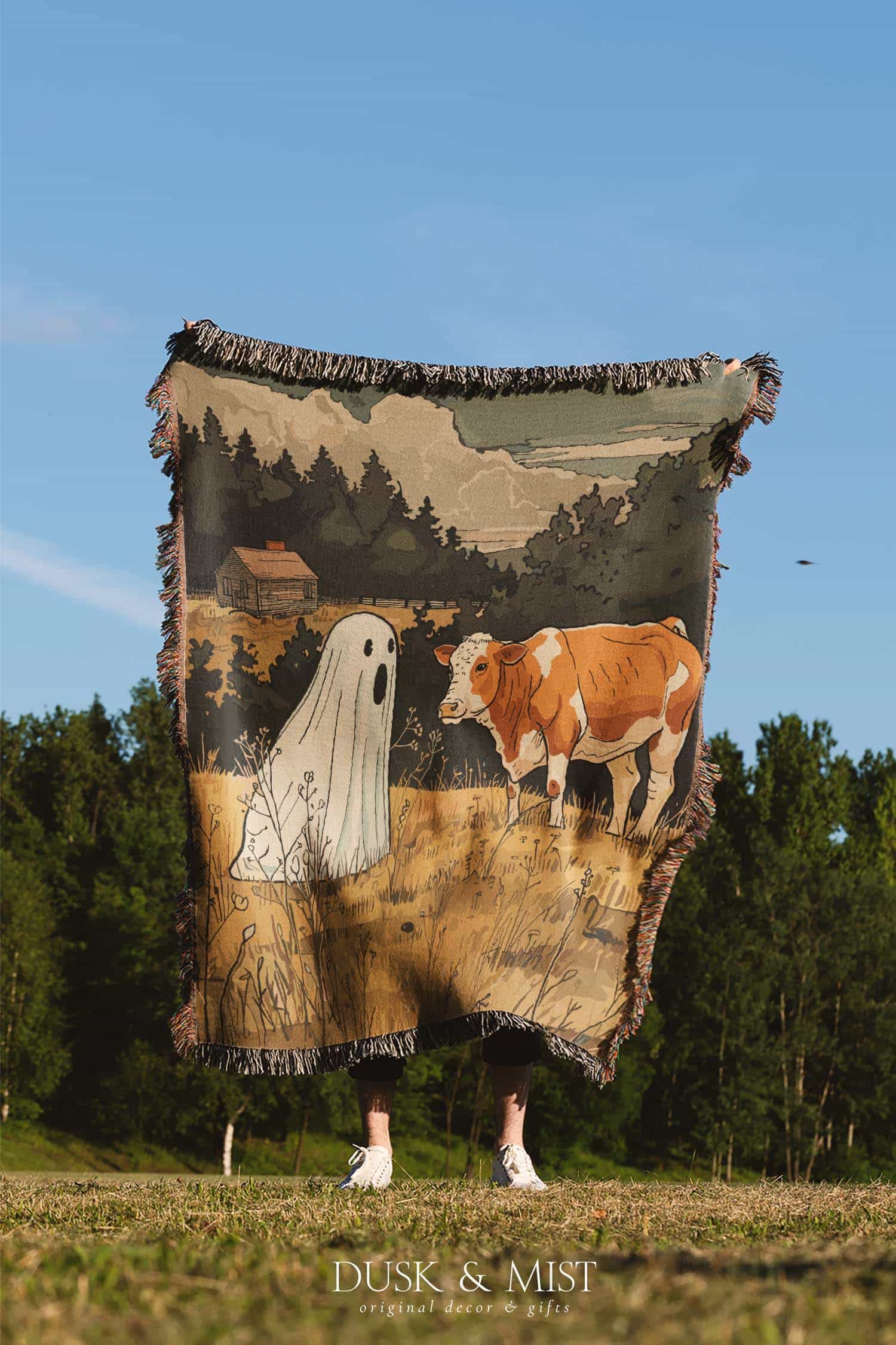 Woven cotton blanket featuring a small ghost standing in a golden meadow facing a brown and white cow, with a rustic cabin and forested hills in the background.