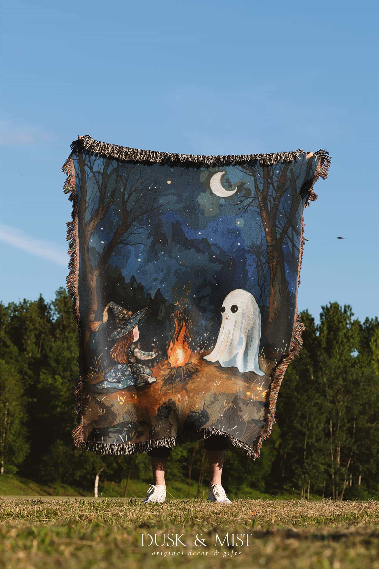 Woven cotton blanket featuring a witch and a ghost sitting by a campfire under a crescent moon in a dark forest setting.