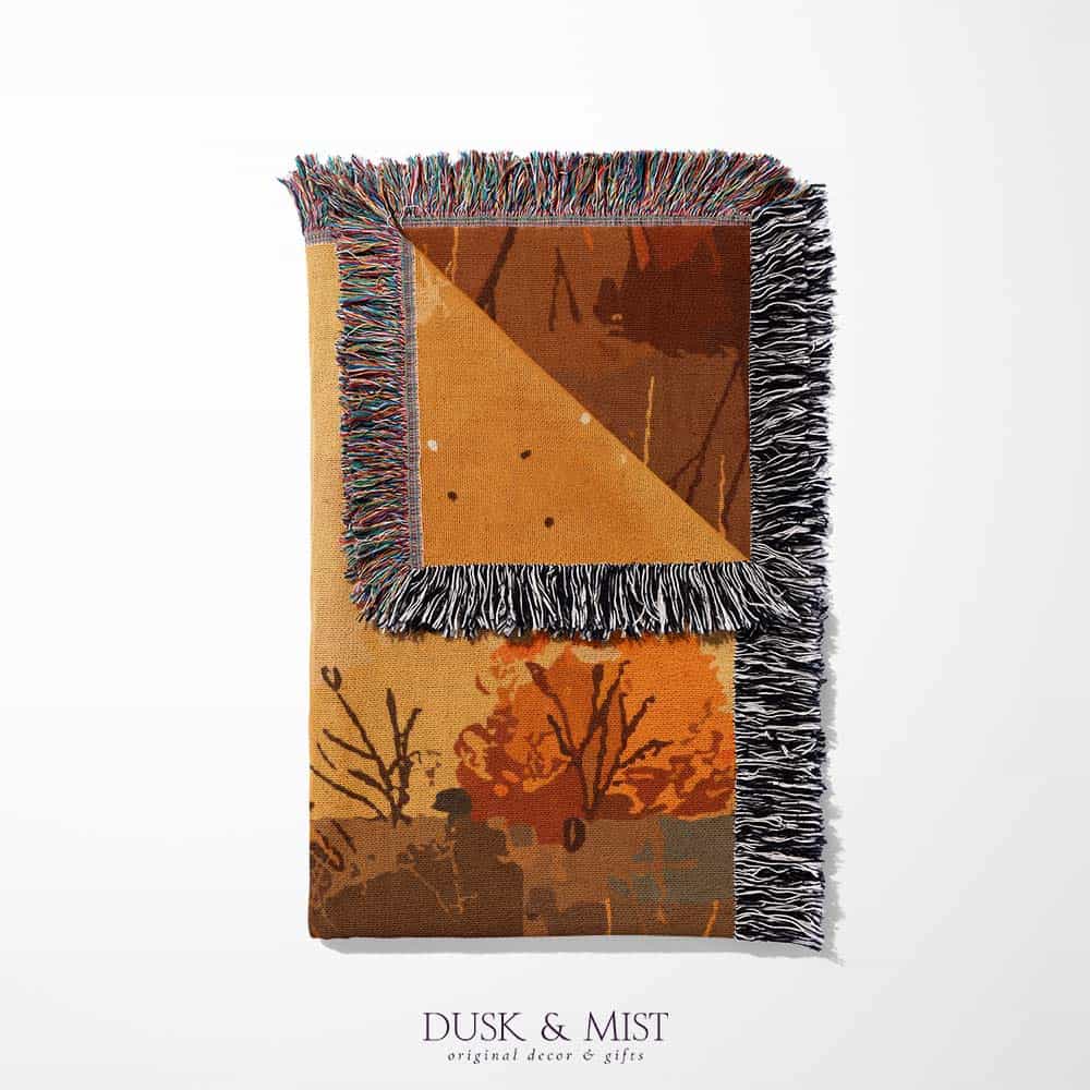 Woven cotton blanket showing a small ghost holding a basket, surrounded by pumpkins and autumn foliage in warm golden and orange tones.
