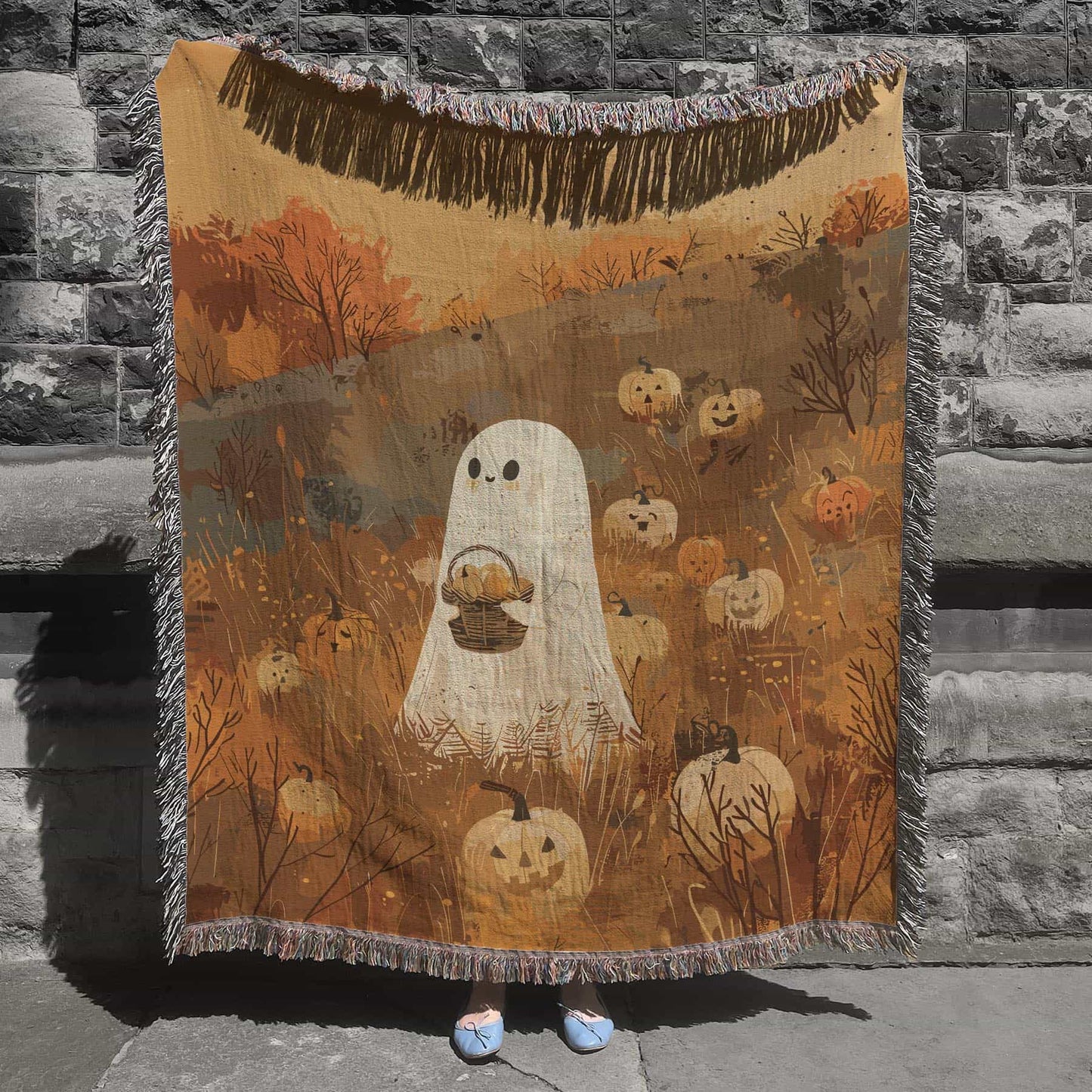 Woven cotton blanket showing a small ghost holding a basket, surrounded by pumpkins and autumn foliage in warm golden and orange tones.
