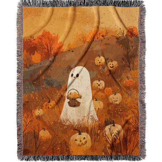 Woven cotton blanket showing a small ghost holding a basket, surrounded by pumpkins and autumn foliage in warm golden and orange tones.