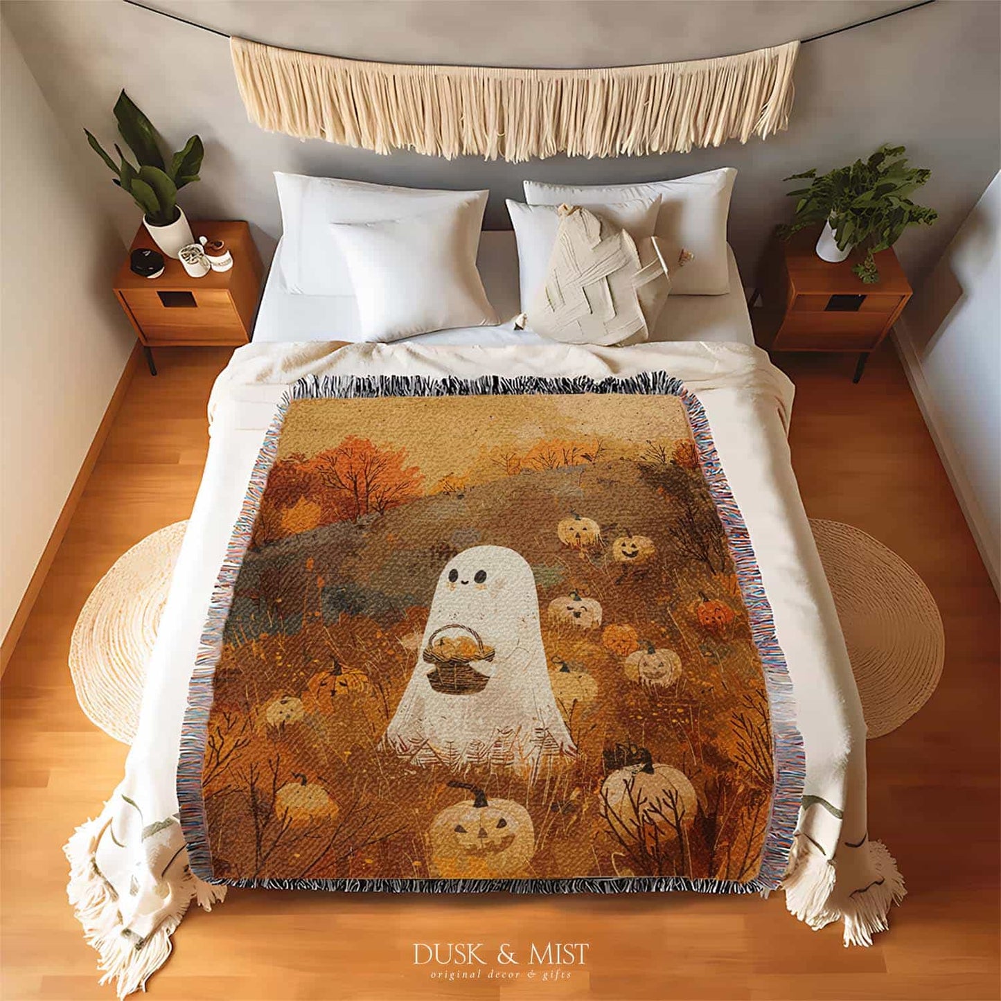 Woven cotton blanket showing a small ghost holding a basket, surrounded by pumpkins and autumn foliage in warm golden and orange tones.