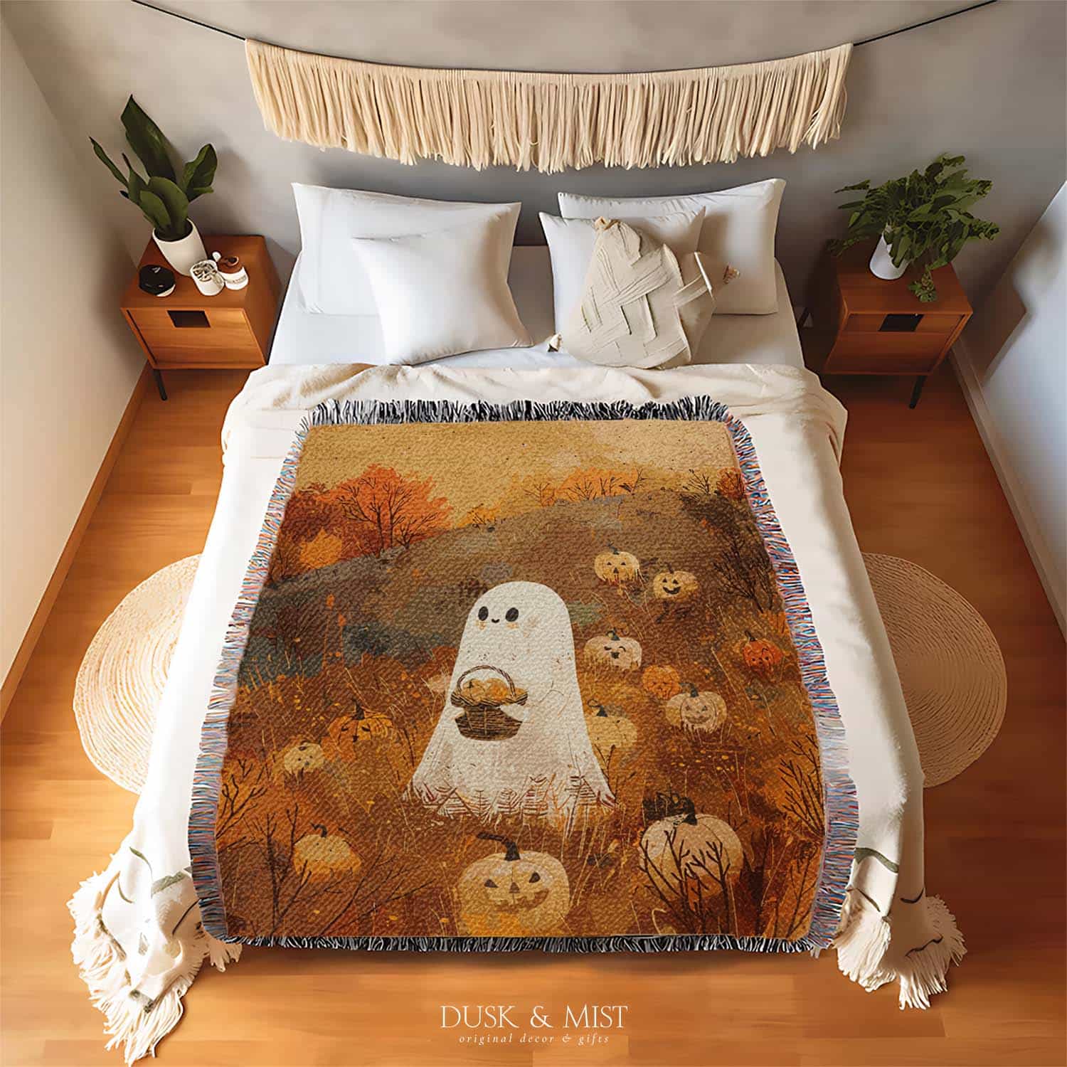 Woven cotton blanket showing a small ghost holding a basket, surrounded by pumpkins and autumn foliage in warm golden and orange tones.
