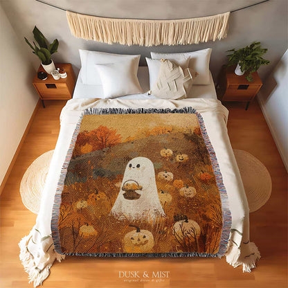Woven cotton blanket showing a small ghost holding a basket, surrounded by pumpkins and autumn foliage in warm golden and orange tones.