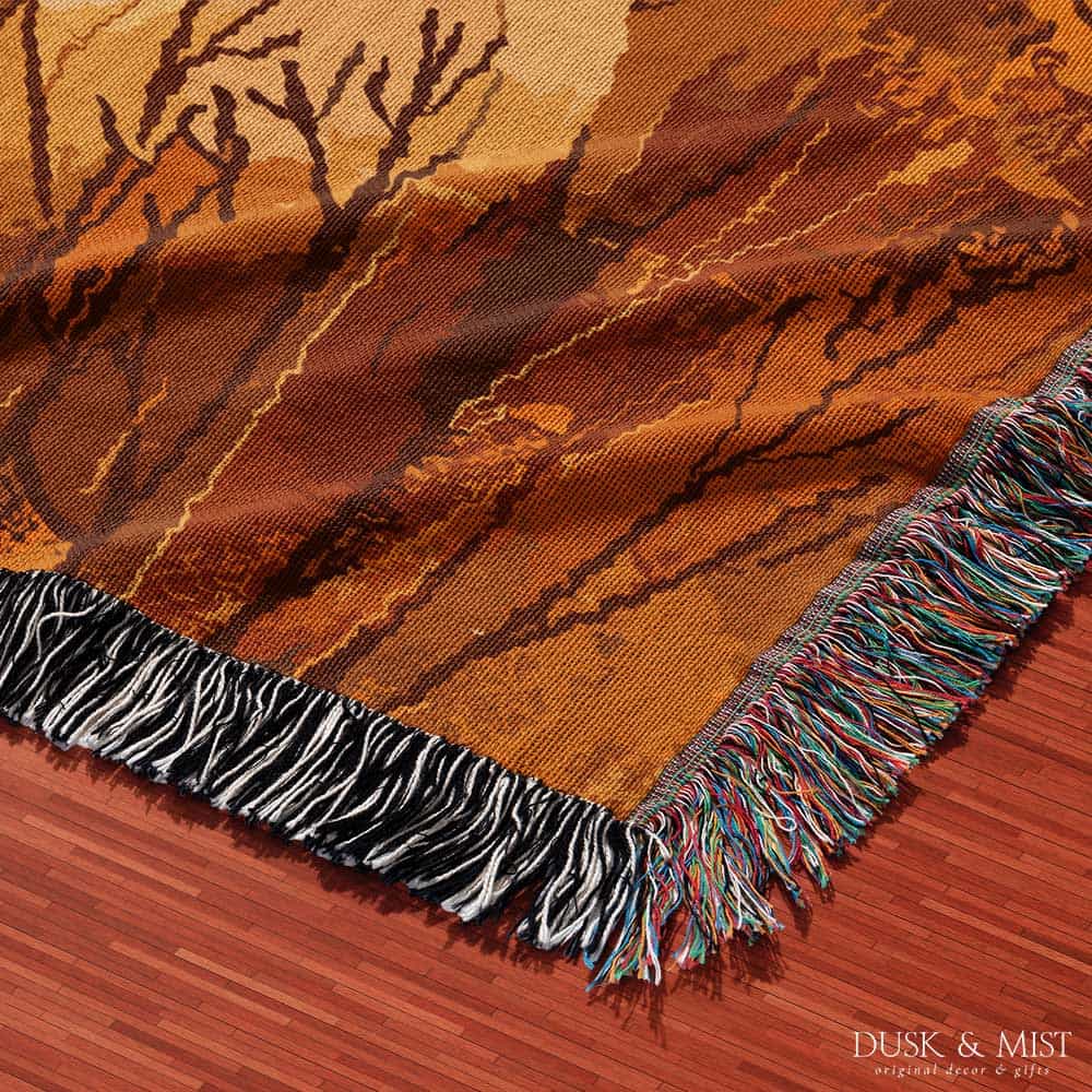 Woven cotton blanket showing a small ghost holding a basket, surrounded by pumpkins and autumn foliage in warm golden and orange tones.