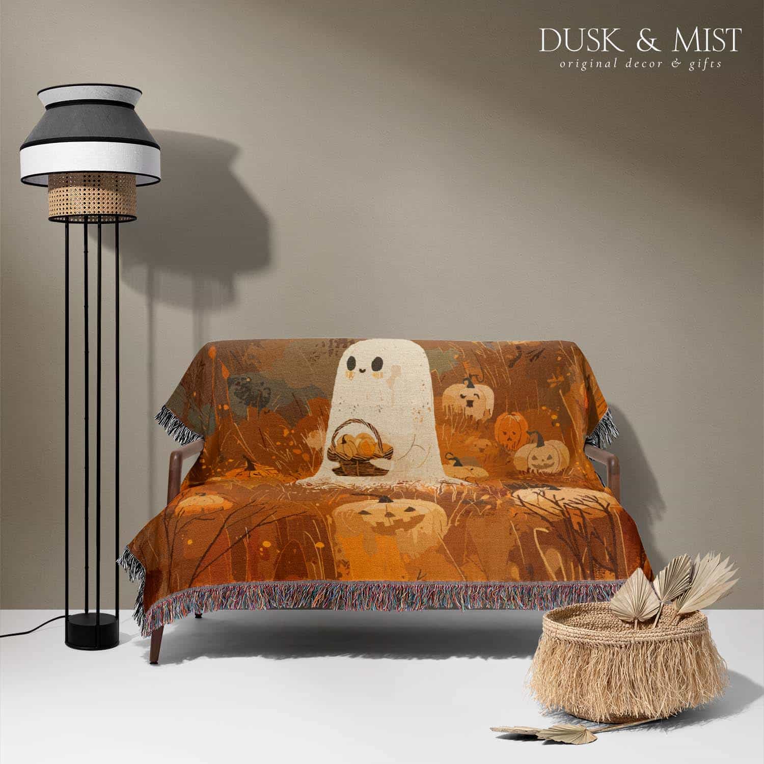 Woven cotton blanket showing a small ghost holding a basket, surrounded by pumpkins and autumn foliage in warm golden and orange tones.