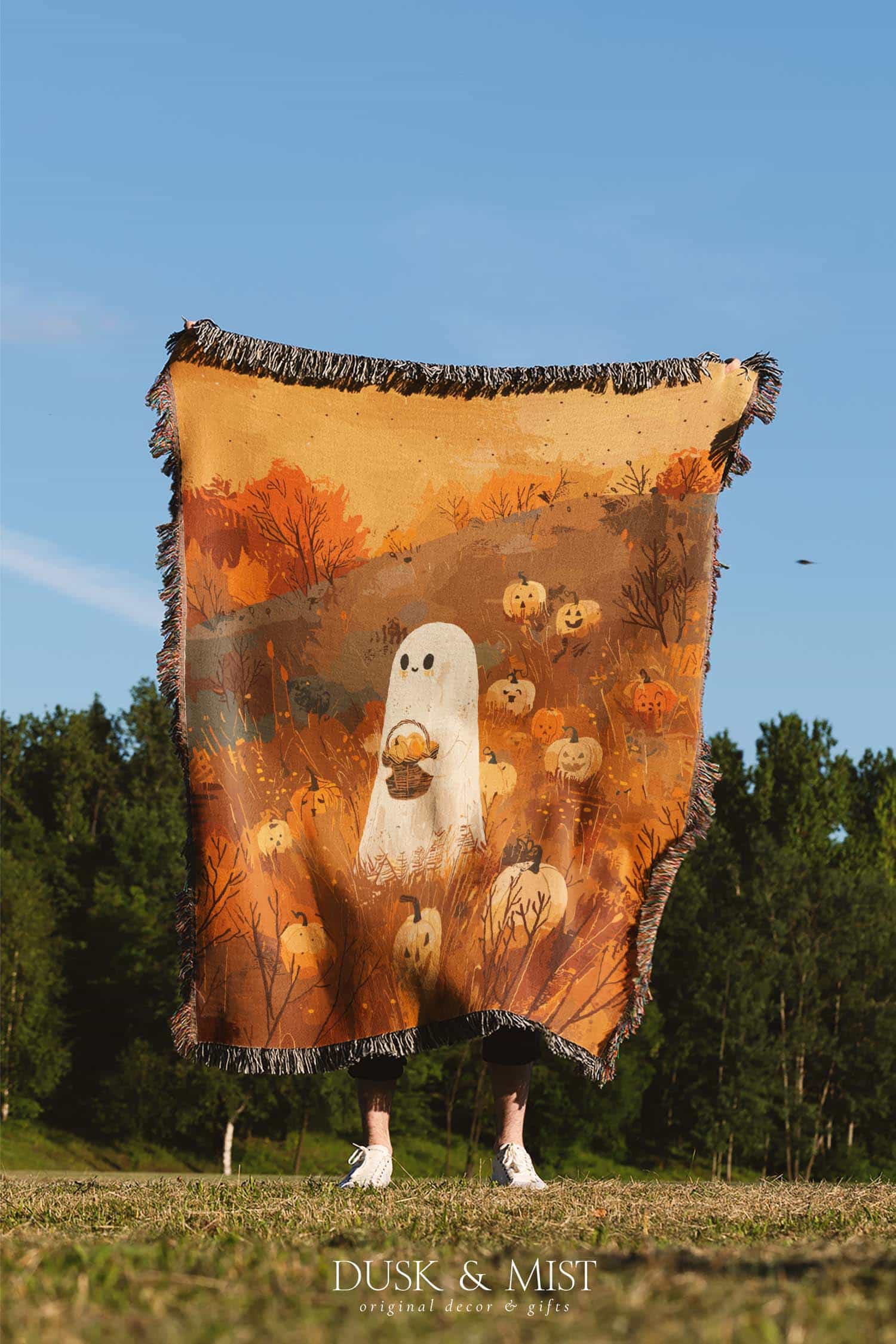 Woven cotton blanket showing a small ghost holding a basket, surrounded by pumpkins and autumn foliage in warm golden and orange tones.