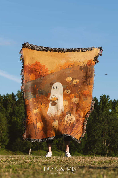Woven cotton blanket showing a small ghost holding a basket, surrounded by pumpkins and autumn foliage in warm golden and orange tones.