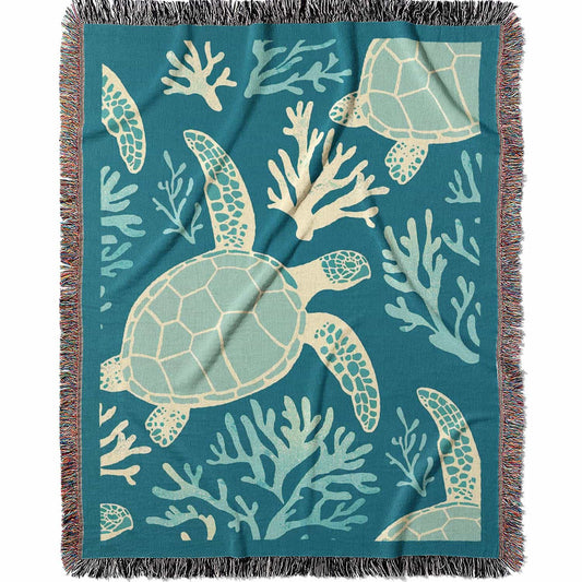 Woven cotton blanket featuring sea turtles swimming among light blue coral on a teal background.  