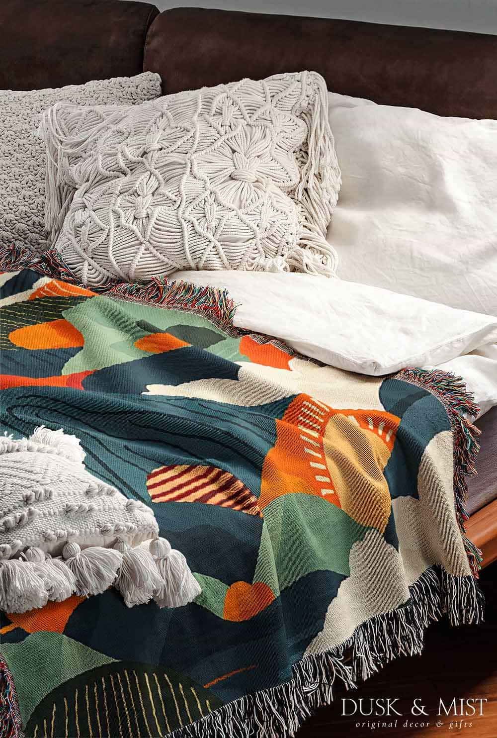Woven cotton blanket featuring an abstract mountain landscape with layered peaks, sun motifs, and bold colors of orange, green, teal, and cream.