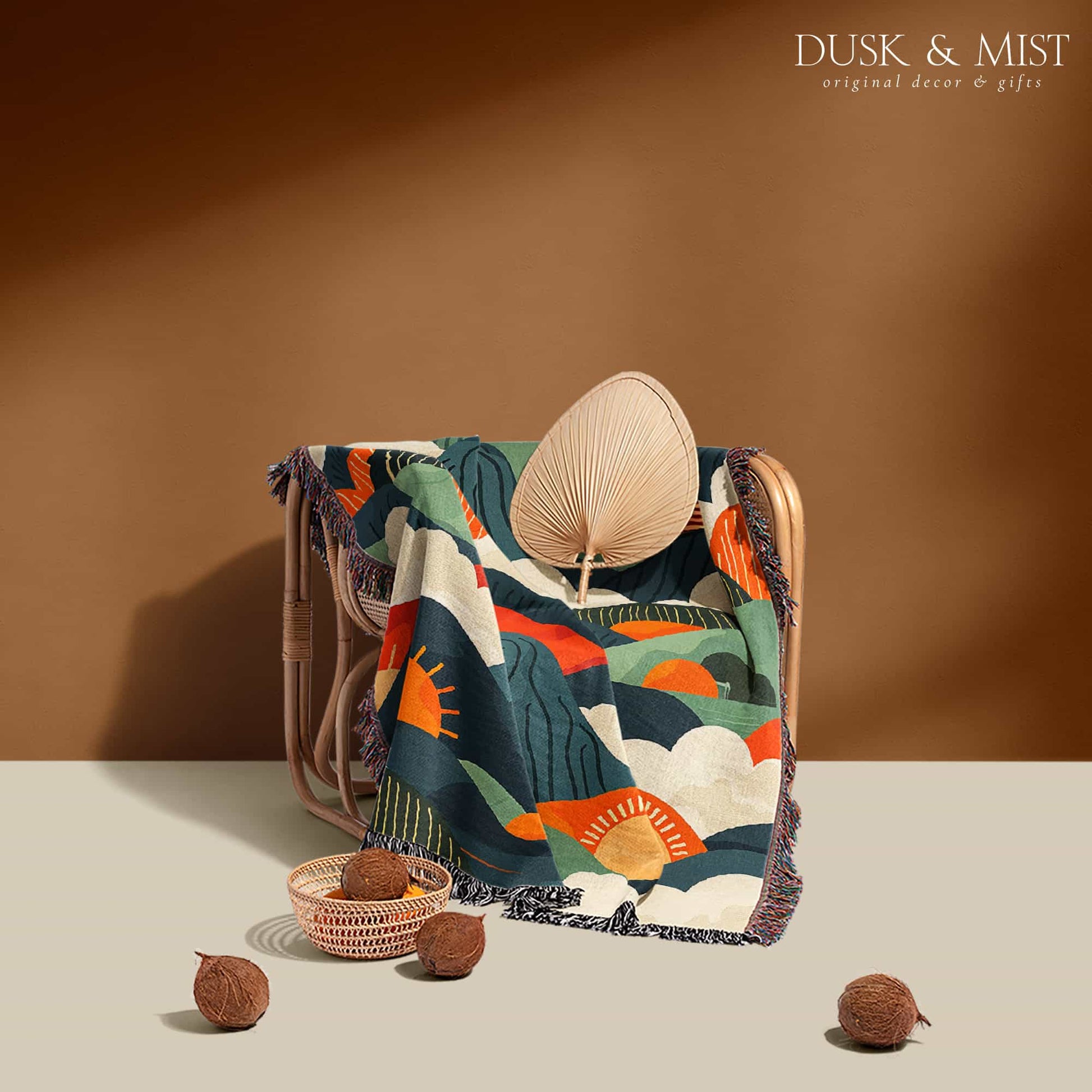 Woven cotton blanket featuring an abstract mountain landscape with layered peaks, sun motifs, and bold colors of orange, green, teal, and cream.