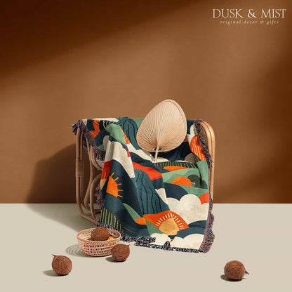 Woven cotton blanket featuring an abstract mountain landscape with layered peaks, sun motifs, and bold colors of orange, green, teal, and cream.
