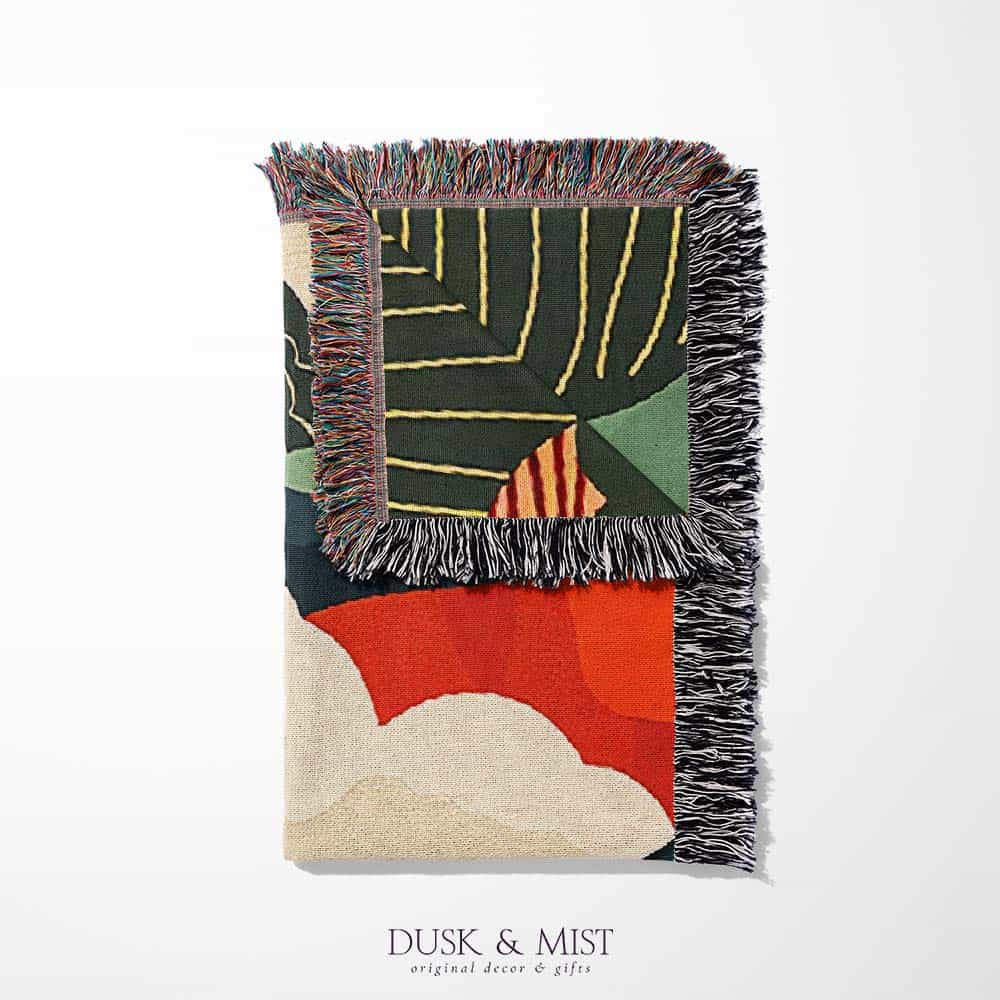 Woven cotton blanket featuring an abstract mountain landscape with layered peaks, sun motifs, and bold colors of orange, green, teal, and cream.