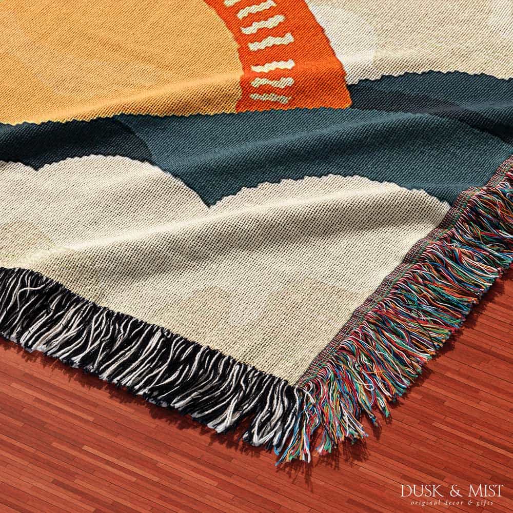 Woven cotton blanket featuring an abstract mountain landscape with layered peaks, sun motifs, and bold colors of orange, green, teal, and cream.