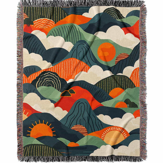 Woven cotton blanket featuring an abstract mountain landscape with layered peaks, sun motifs, and bold colors of orange, green, teal, and cream.
