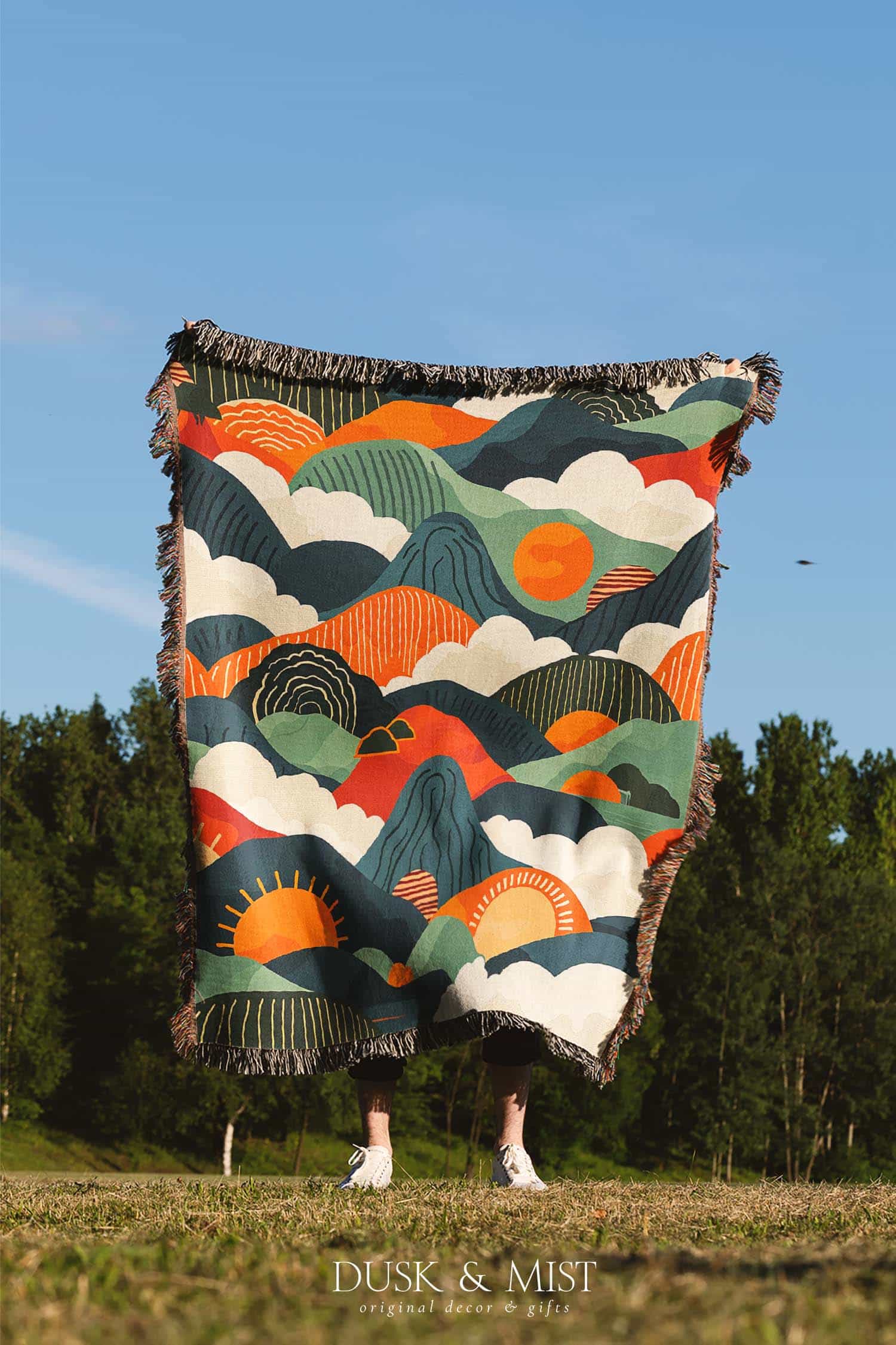 Woven cotton blanket featuring an abstract mountain landscape with layered peaks, sun motifs, and bold colors of orange, green, teal, and cream.