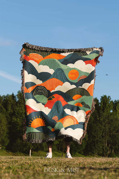 Woven cotton blanket featuring an abstract mountain landscape with layered peaks, sun motifs, and bold colors of orange, green, teal, and cream.
