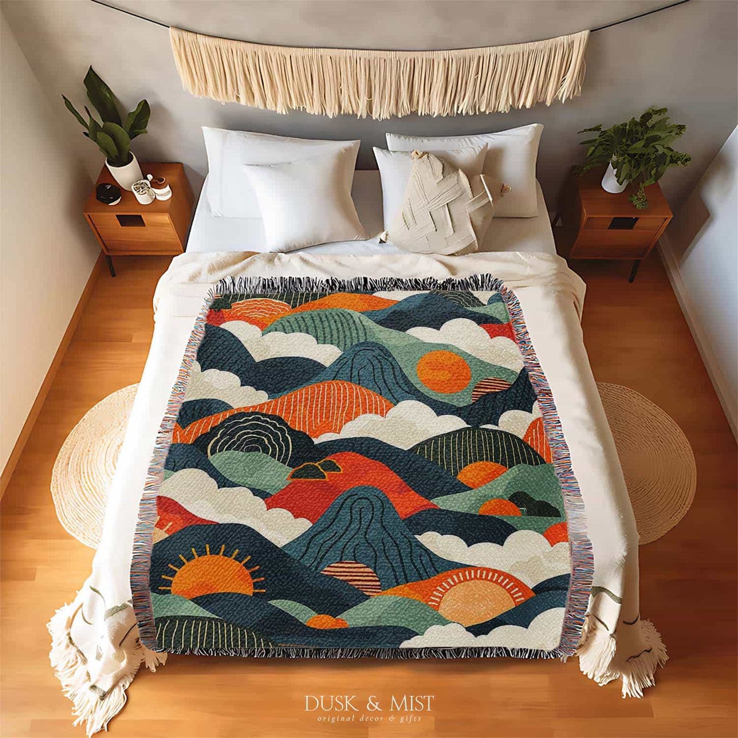 Woven cotton blanket featuring an abstract mountain landscape with layered peaks, sun motifs, and bold colors of orange, green, teal, and cream.