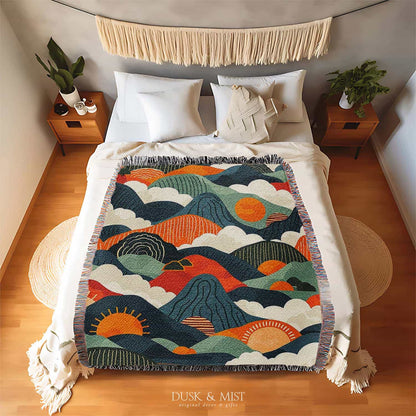 Woven cotton blanket featuring an abstract mountain landscape with layered peaks, sun motifs, and bold colors of orange, green, teal, and cream.