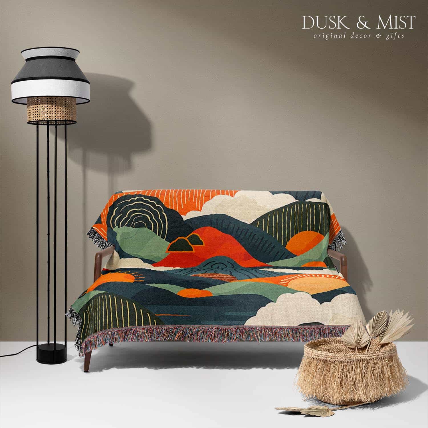 Woven cotton blanket featuring an abstract mountain landscape with layered peaks, sun motifs, and bold colors of orange, green, teal, and cream.