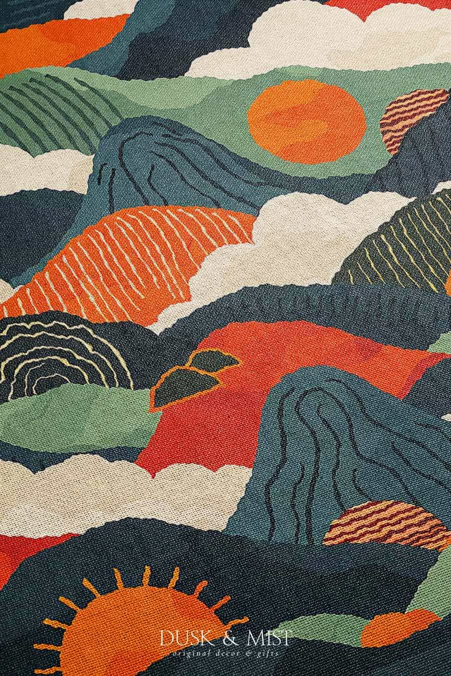 Woven cotton blanket featuring an abstract mountain landscape with layered peaks, sun motifs, and bold colors of orange, green, teal, and cream.