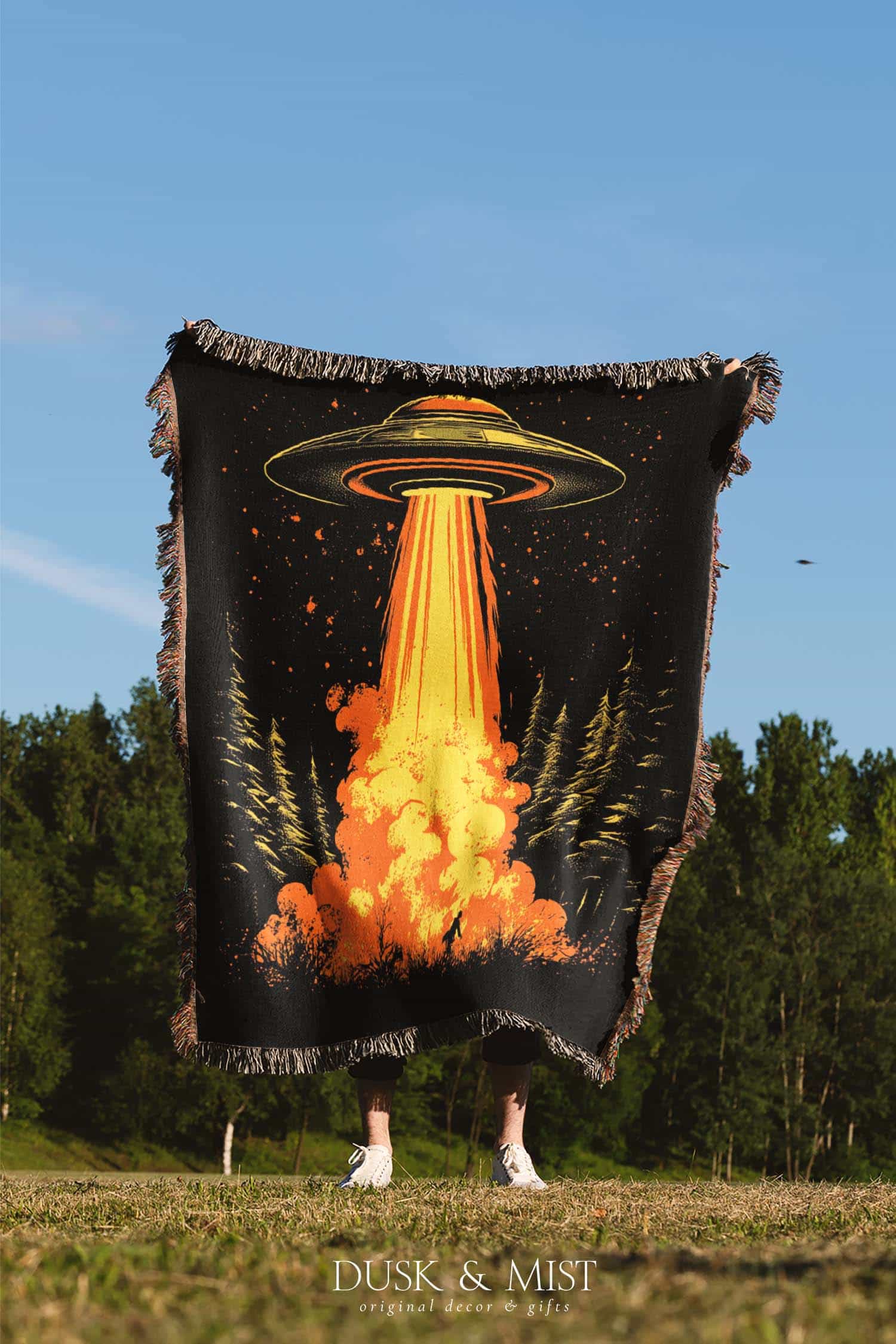 Woven cotton blanket featuring a UFO with a fiery orange beam blasting into a forest, with smoke clouds and a small human silhouette running at the bottom.