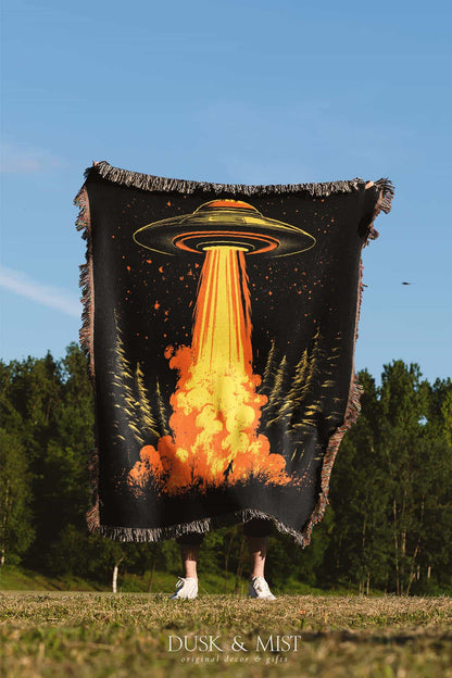 Woven cotton blanket featuring a UFO with a fiery orange beam blasting into a forest, with smoke clouds and a small human silhouette running at the bottom.