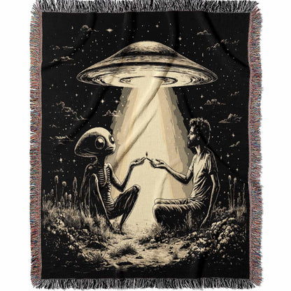 Woven cotton blanket featuring an alien and a human seated under a UFO’s glowing beam, reaching hands toward each other against a starry night sky.
