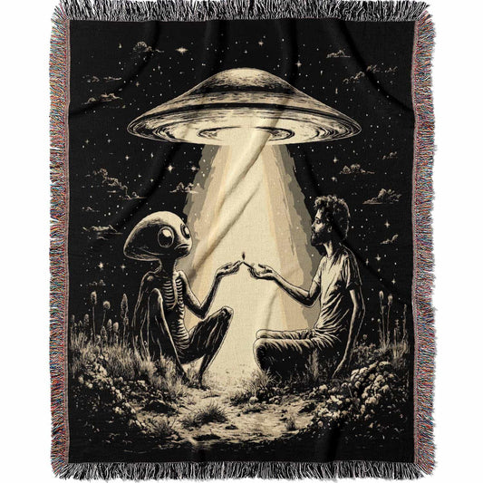 Woven cotton blanket featuring an alien and a human seated under a UFO’s glowing beam, reaching hands toward each other against a starry night sky.
