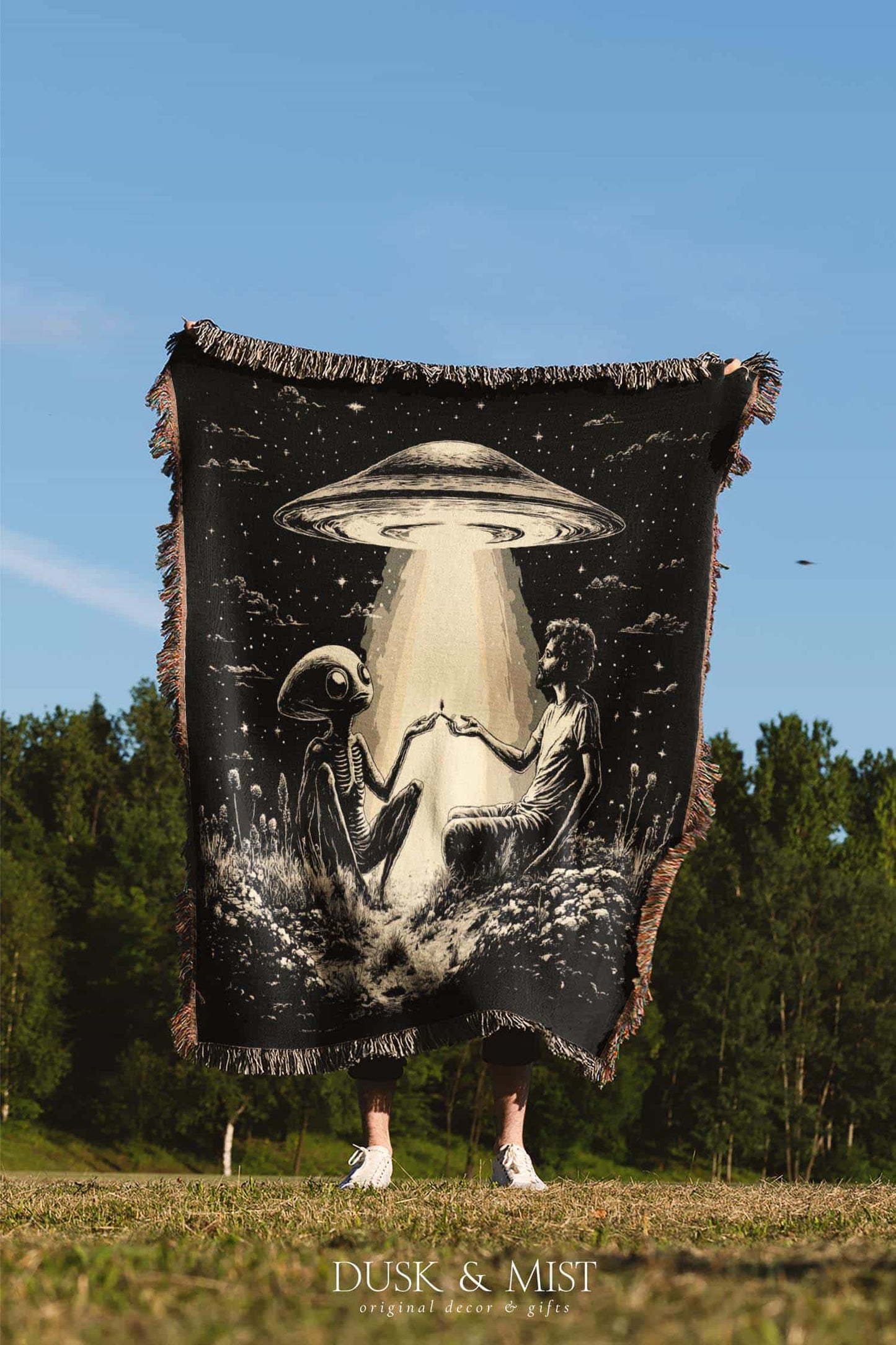 Woven cotton blanket featuring an alien and a human seated under a UFO’s glowing beam, reaching hands toward each other against a starry night sky.