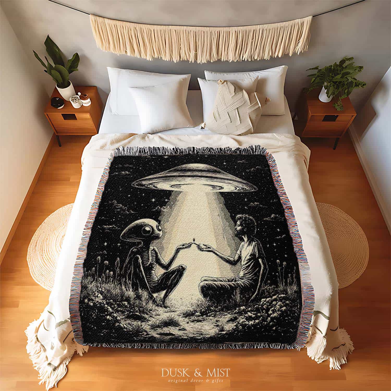 Woven cotton blanket featuring an alien and a human seated under a UFO’s glowing beam, reaching hands toward each other against a starry night sky.