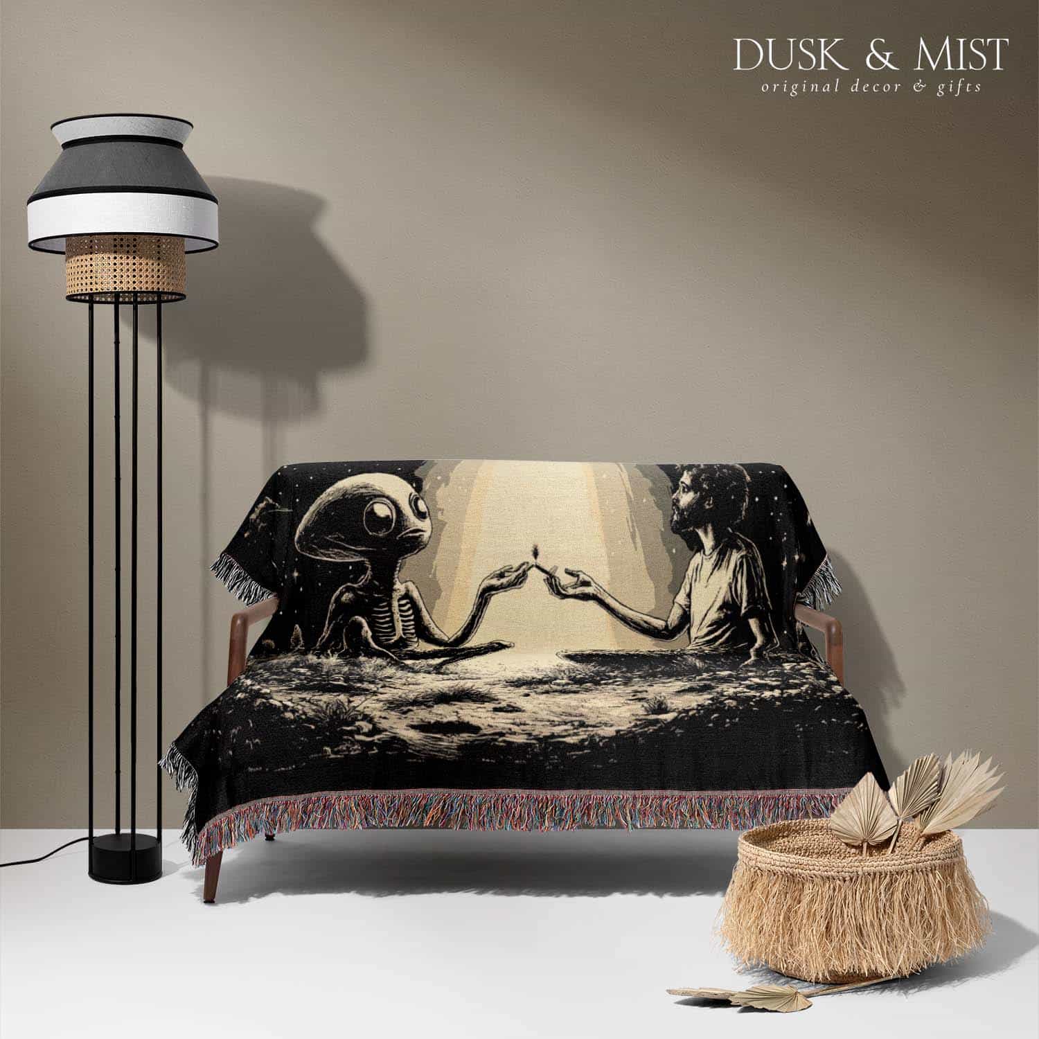 Woven cotton blanket featuring an alien and a human seated under a UFO’s glowing beam, reaching hands toward each other against a starry night sky.