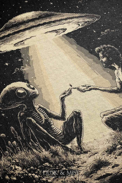 Woven cotton blanket featuring an alien and a human seated under a UFO’s glowing beam, reaching hands toward each other against a starry night sky.