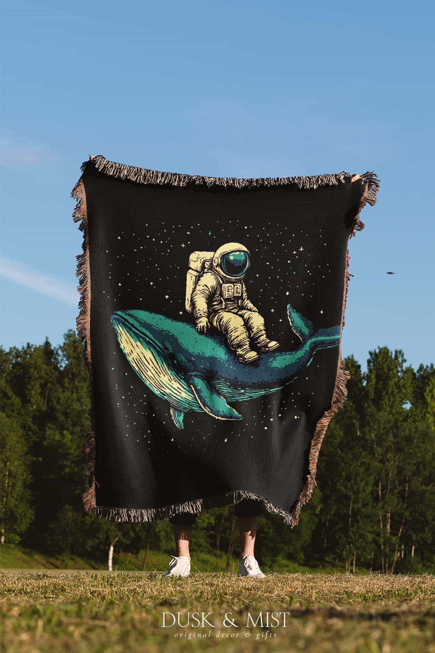 Woven cotton blanket featuring an astronaut riding a whale through space with colorful stars on a black background.