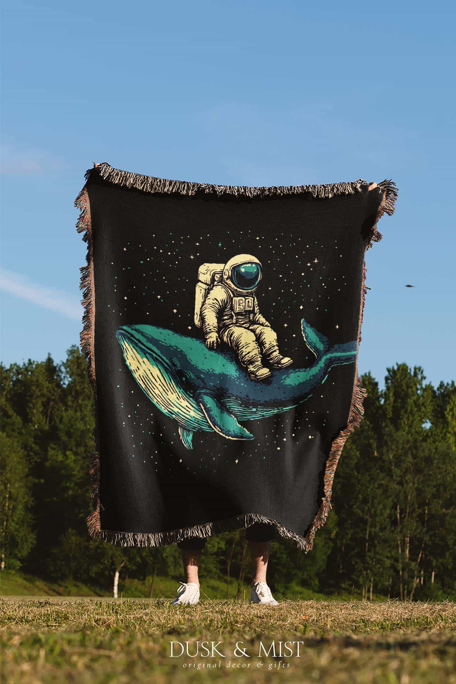 Woven cotton blanket featuring an astronaut riding a whale through space with colorful stars on a black background.