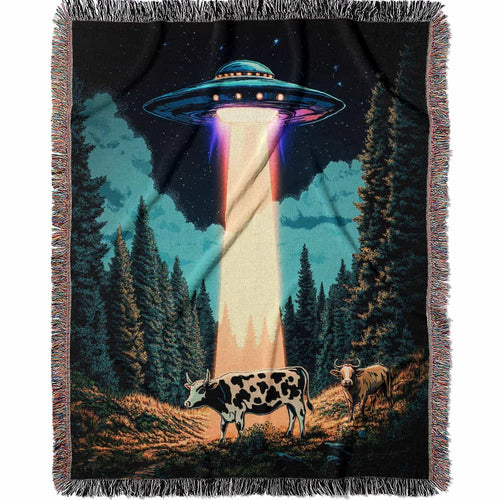 Woven cotton blanket showing a UFO beaming light onto a cow in a forest clearing under a starry night sky with trees and clouds.