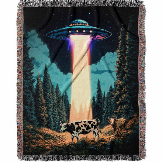 Woven cotton blanket showing a UFO beaming light onto a cow in a forest clearing under a starry night sky with trees and clouds.