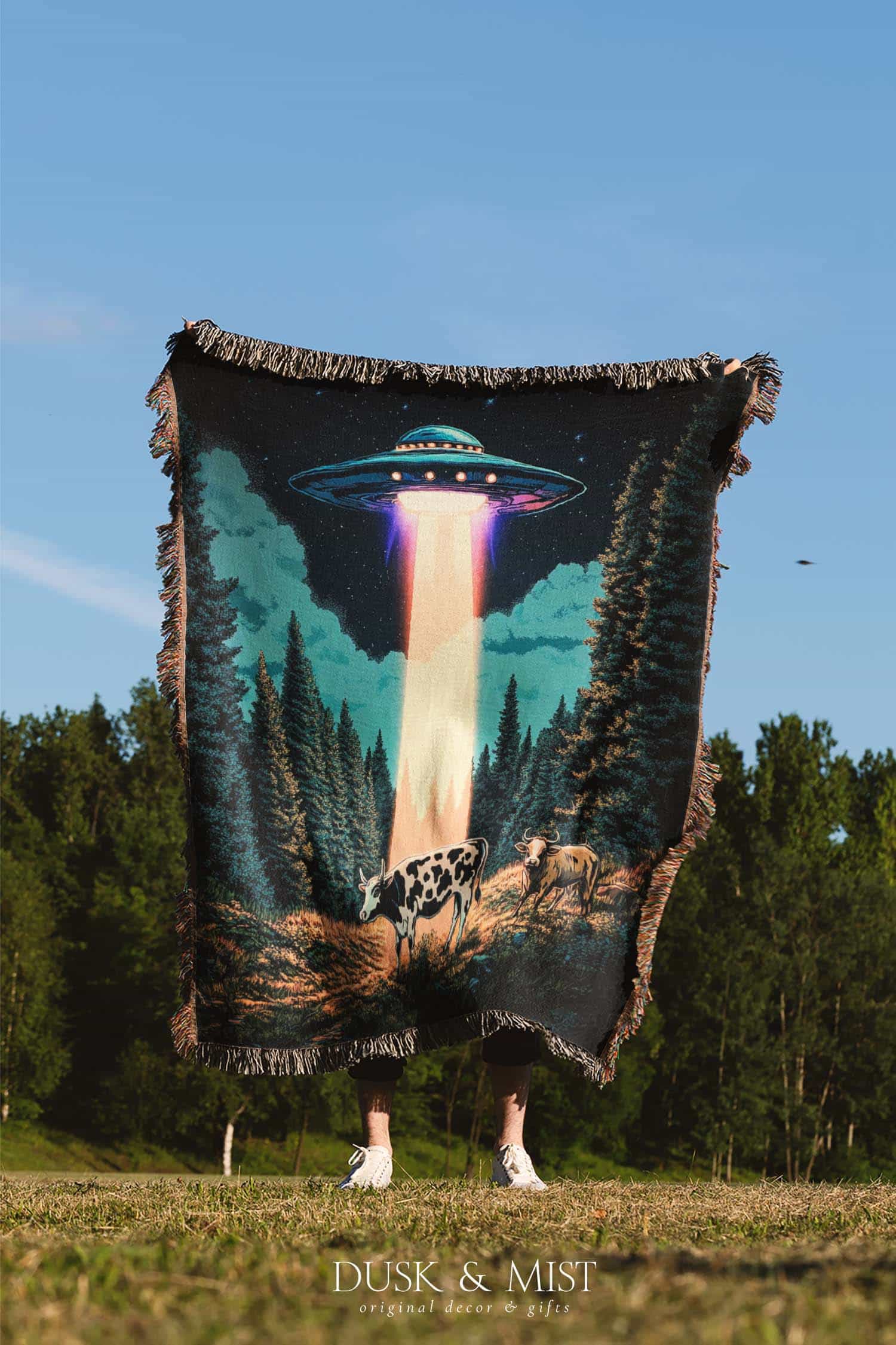 Woven cotton blanket showing a UFO beaming light onto a cow in a forest clearing under a starry night sky with trees and clouds.