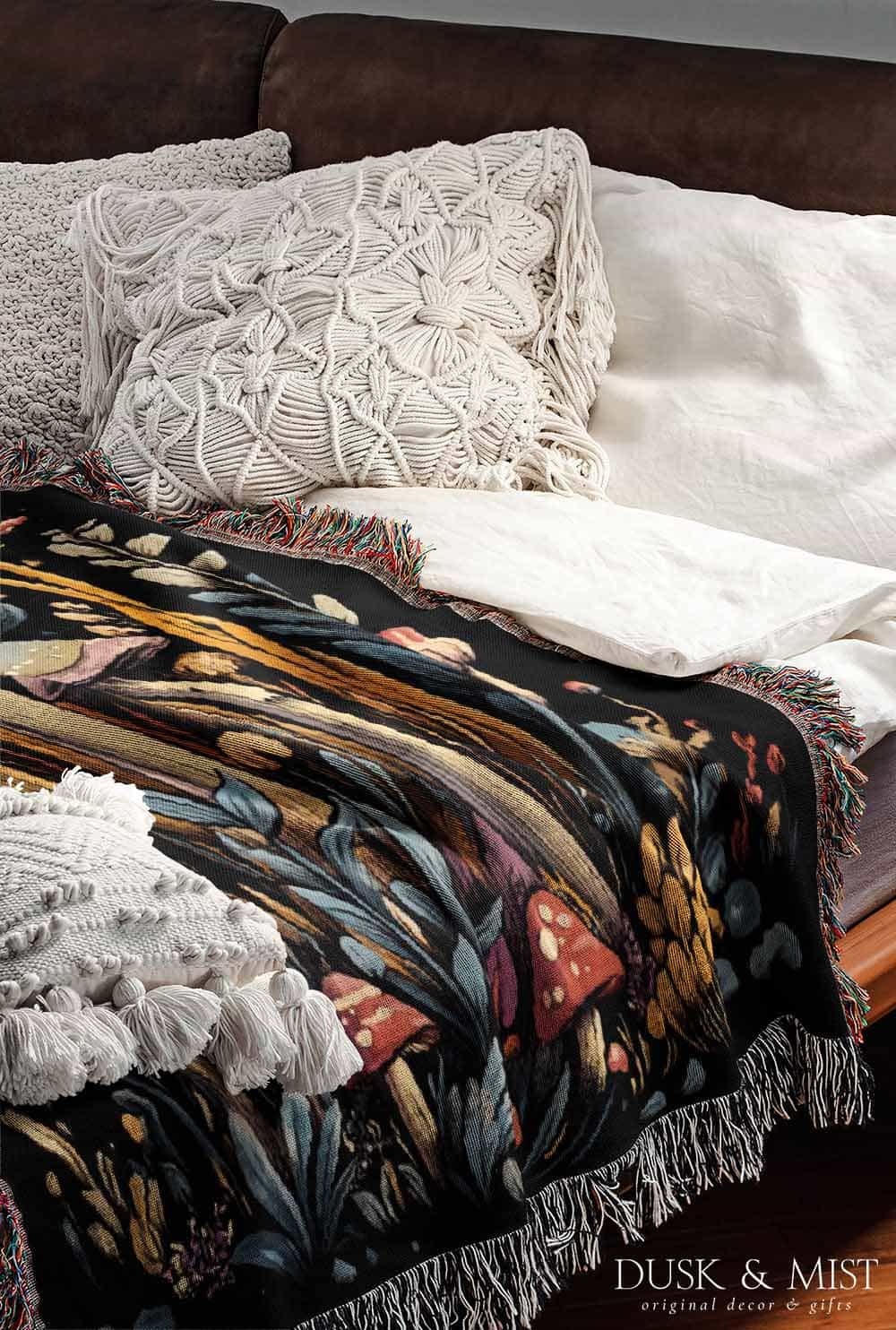 Woven cotton blanket featuring a botanical-style cluster of mushrooms in red, gold, and purple caps surrounded by plants and foliage on a black background.