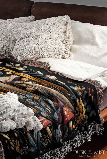 Woven cotton blanket featuring a botanical-style cluster of mushrooms in red, gold, and purple caps surrounded by plants and foliage on a black background.