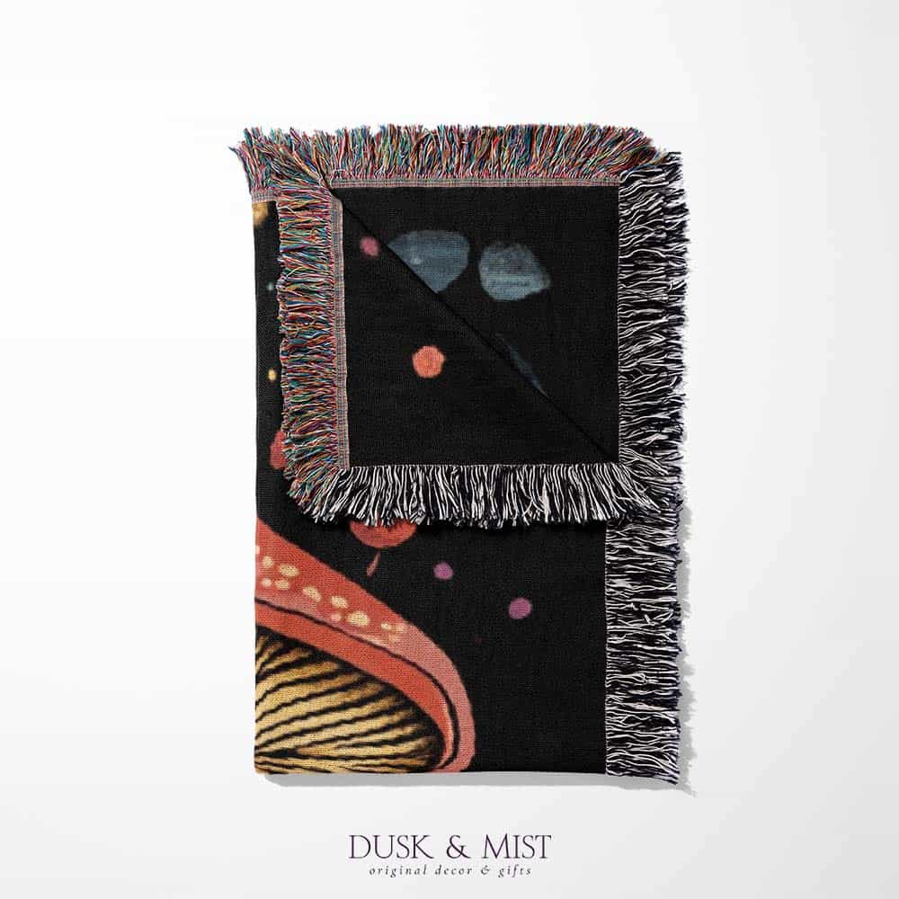 Woven cotton blanket featuring a botanical-style cluster of mushrooms in red, gold, and purple caps surrounded by plants and foliage on a black background.